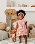 Bébé debout portant l’ensemble robe imprimée Bandia de Bobo Bassari, avec une poupée assortie aux motifs africains roses