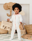 Bébé debout portant le pyjama bébé été col imprimé Bandia de Bobo Bassari, tenant une maisonnette en rotin dans une main et une pochette en rotin dans l'autre, illustrant une ambiance naturelle et ludique