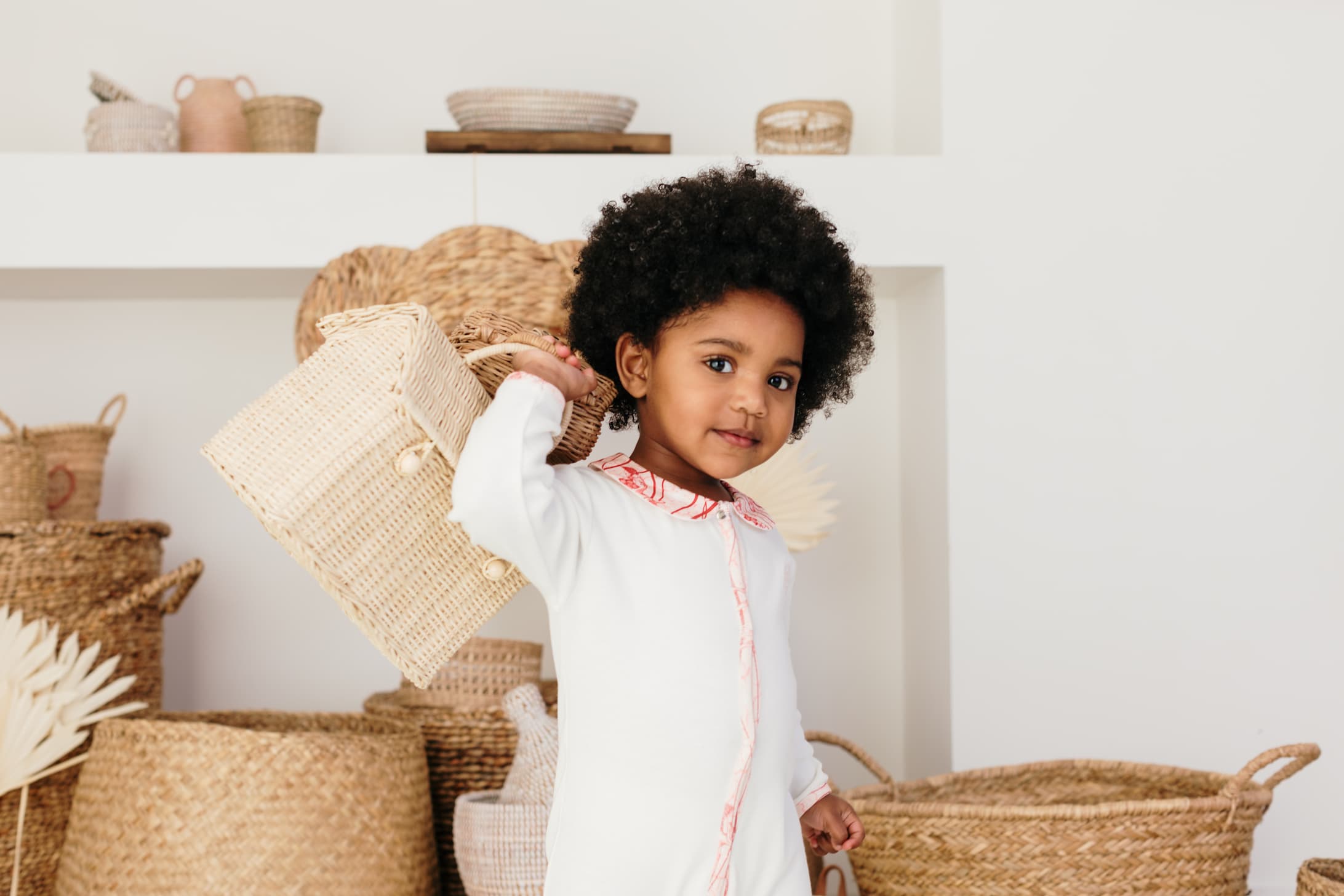 Bébé debout portant le pyjama bébé été col imprimé Bandia de Bobo Bassari, tenant une maisonnette en rotin dans une main et une pochette en rotin dans l'autre, illustrant une ambiance naturelle et ludique