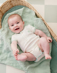 Un bébé tout sourire allongé dans un panier en rosier portant le Body bébé Samé de la marque Bobo Bassari, avec un design inspiré de motifs africains, en coton doux, adapté pour les nouveaux-nés.