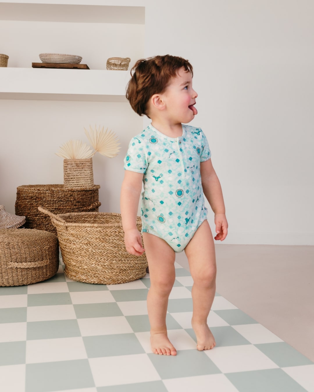 Bébé debout portant le corps imprimé Samé de Bobo Bassari, regardant vers sa gauche et tirant la langue, capturant un moment ludique et naturel