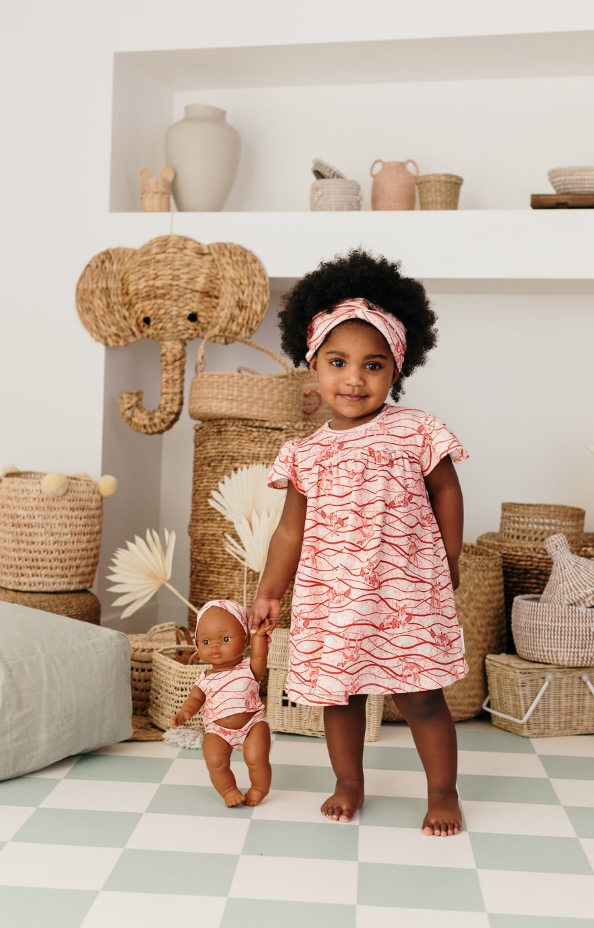 Bébé debout portant l’ensemble robe imprimée Bandia de Bobo Bassari, avec une poupée assortie aux motifs africains roses